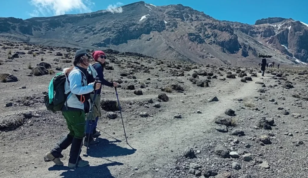 Two hikers with trekking poles and backpacks traverse a rocky volcanic landscape toward rugged mountains under a clear blue sky