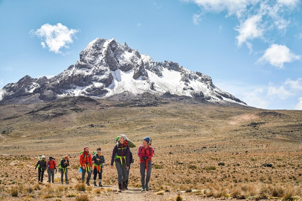 Group of hikers in colorful jackets walking across a rocky plain with a snowcapped mountain in the background across a blue sky