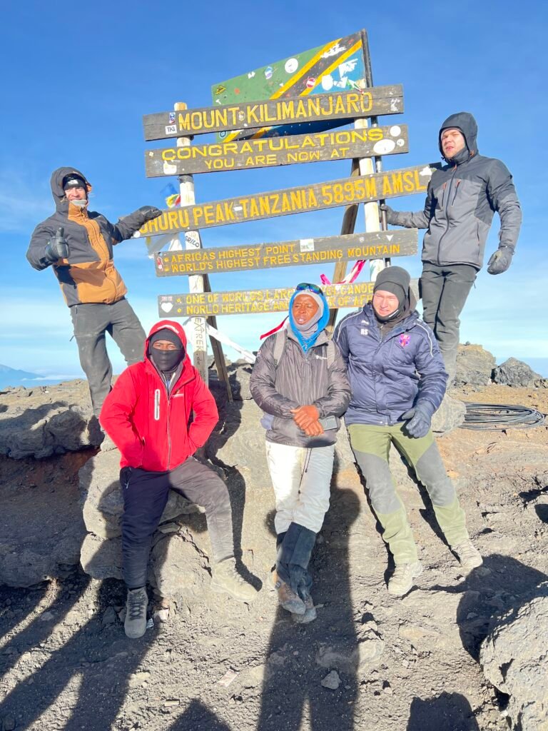 Group of Climbers Posing at the Mount Kilimanjaro Summit Sign Against a Clear Blue Sky with Elevation Text Visible uhuru Peak 5895 Mgroup of Hikers at Kilimanjaro Summit Sign with Blue Sky Behind Them Wearing Winter Jackets and Gloves Looking at Camera