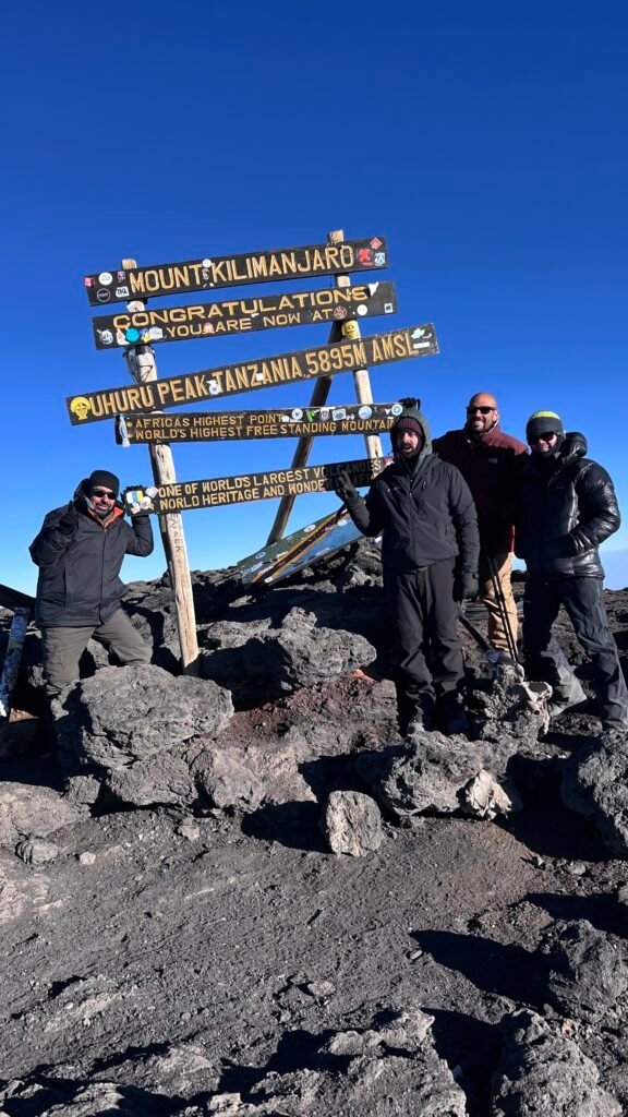 Group of Climbers Posing Beside a Wooden Kilimanjaro Summit Sign 5895 M Above Sea Level on a Rocky Volcanic Terrain Under a Bright Blue Sky