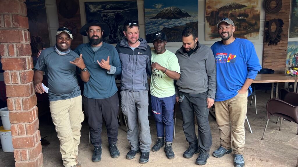 Six Men Stand Together Indoors Smiling for the Camera Wearing Casual Outdoor Gear and Hiking Boots