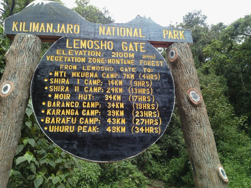 Kilimanjaro National Park Signboard with Yellow Lettering in a Forest Lists Lemosho Gate and Various Camps with Distances and Times